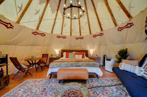 Cozy yurt interior with a bed, seating area, and rustic decor, featuring a chandelier and a stone fire pit.